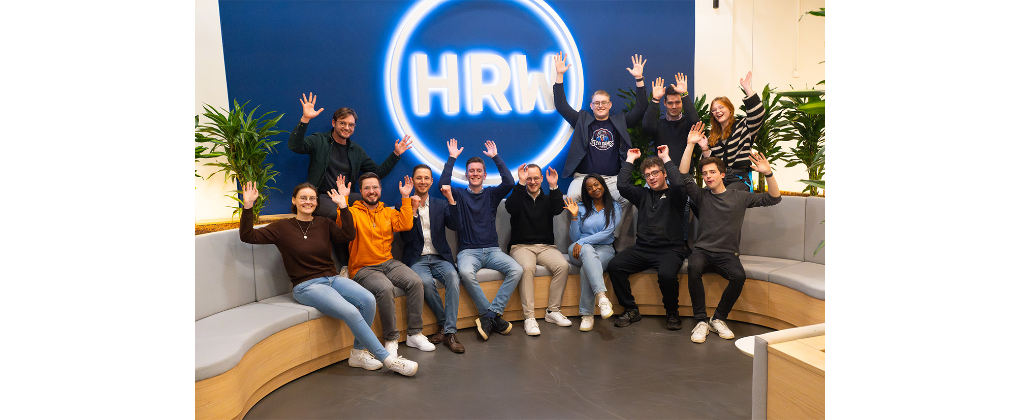 Gruppenfoto von Studierenden und Mitarbeitenden im HRW-Startups-Bereich, die gemeinsam sitzend die Arme hochheben.