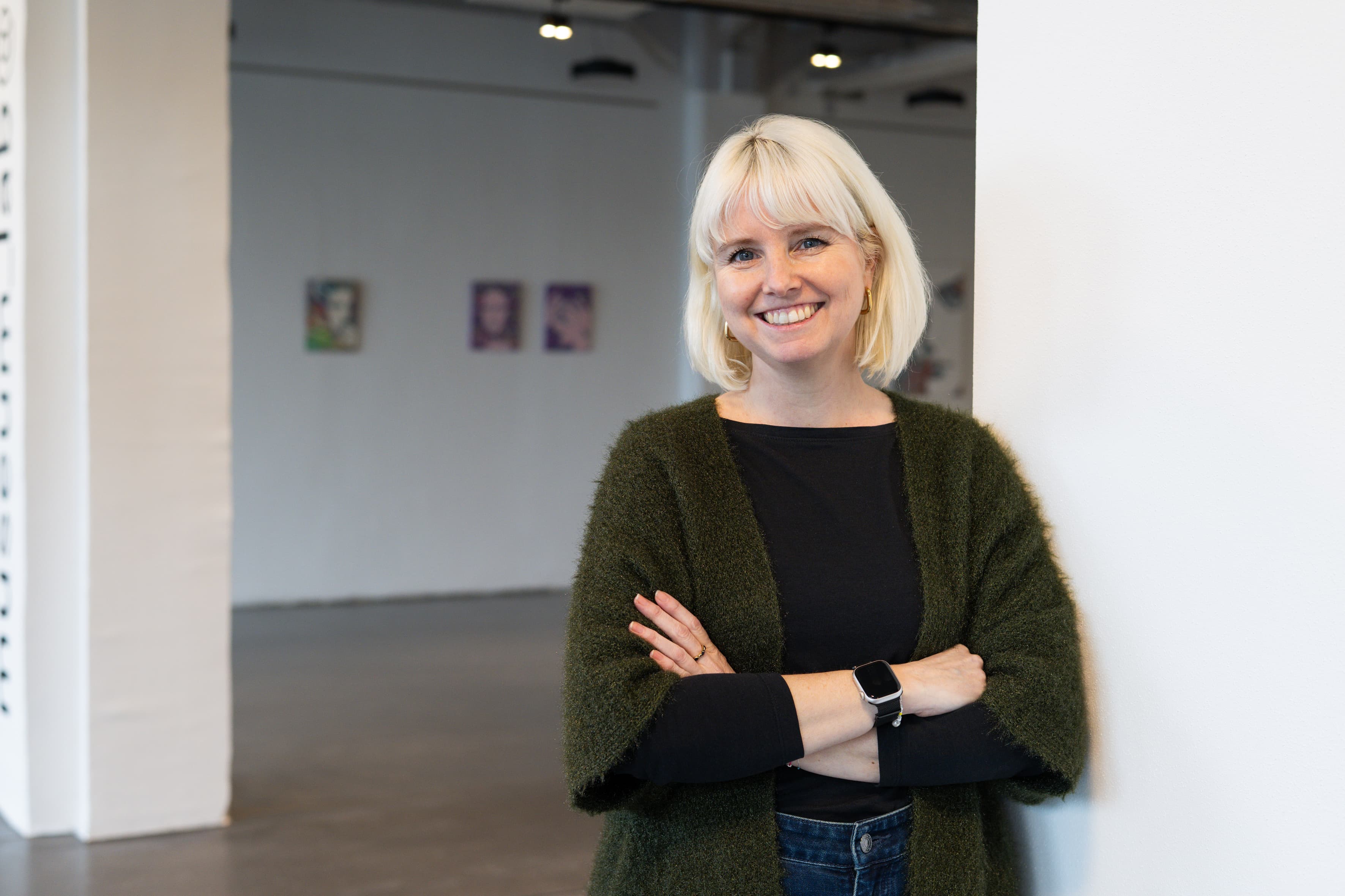 Lächelnde Person mit blondem Bob lehnt an weißer Wand in Galerie; grüner Cardigan, Smartwatch, unscharfe Porträts im Hintergrund.