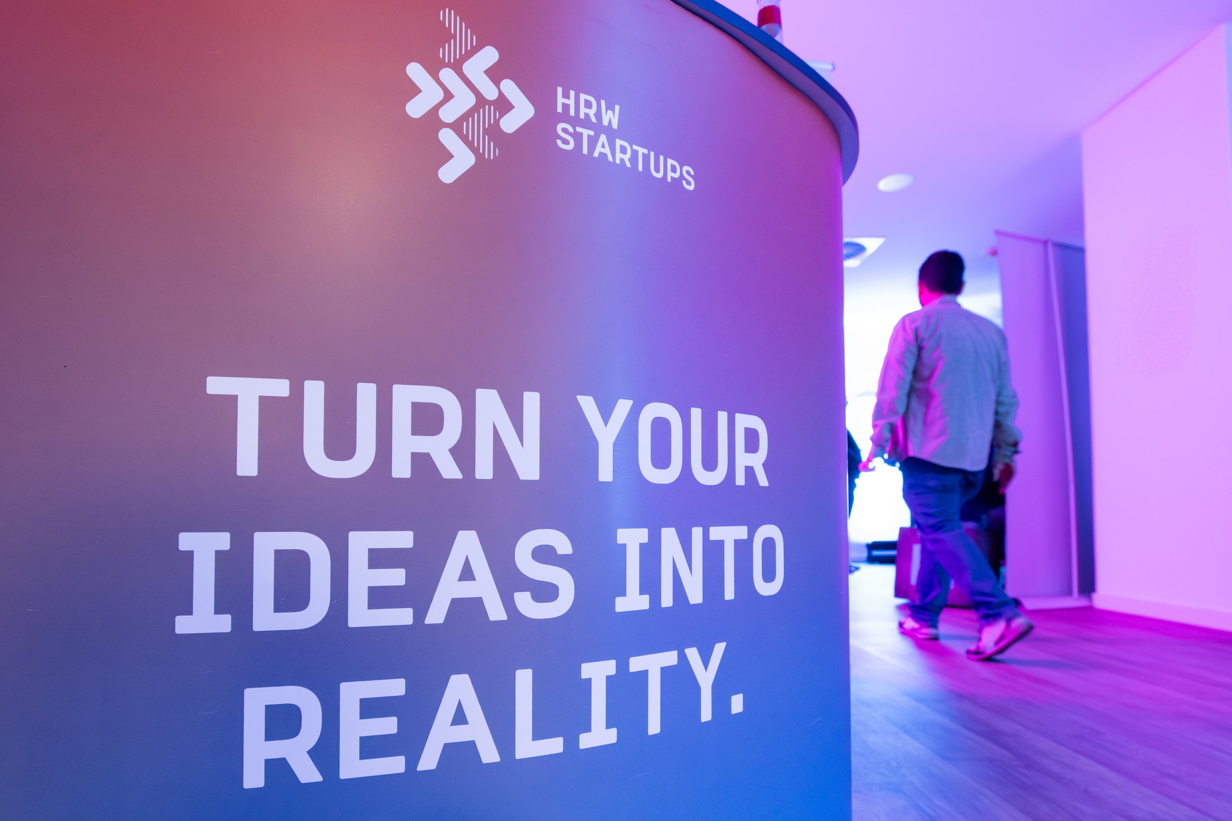 Gebogene Wand mit Logo 'HRW Startups' und Slogan 'TURN YOUR IDEAS INTO REALITY' in pink-blauem Licht; Personen gehen vorbei.